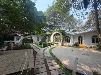 A paved courtyard with a winding stone path leads toward a white building with arched doorways and windows, partially covered in climbing vines. Trees with dense green foliage frame the scene, casting dappled shade over the walkway and surrounding grassy areas. To the left, a tiled patio holds a white metal chair, while a low rope barrier runs along the edge of the path. The building features multiple arched openings, including a central entrance and side windows, with a small yellow step visible at one doorway.