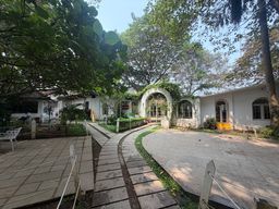 A paved courtyard with a winding stone path leads toward a white building with arched doorways and windows, partially covered in climbing vines. Trees with dense green foliage frame the scene, casting dappled shade over the walkway and surrounding grassy areas. To the left, a tiled patio holds a white metal chair, while a low rope barrier runs along the edge of the path. The building features multiple arched openings, including a central entrance and side windows, with a small yellow step visible at one doorway.