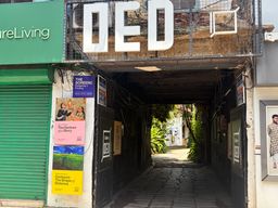 A narrow paved alleyway passes under a building entrance, with large white letters mounted above the opening. To the left, a green storefront with a closed metal shutter stands beside a wall covered in several posters, including one with a pink background and another with a yellow background. To the right, a framed poster shows a person in a patterned shirt. The alley leads toward a brighter area with green foliage visible at the far end.