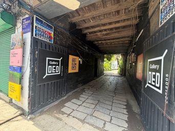A narrow paved alleyway runs between dark walls under a wooden ceiling, with signs and posters attached to the surfaces on both sides and an opening at the far end leading to greenery.