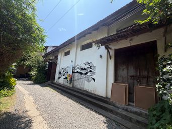 A narrow gravel path runs alongside a long white building under a bright sky, with green foliage on both sides. The building has a dark tiled roof and a large wooden door set into the wall, with two brown panels leaning against the steps in front of it. Along the side of the building, a black-and-white mural covers part of the wall, and a vertical sign stands near the artwork. Small windows are visible above the mural, and power lines stretch across the sky above the roofline.