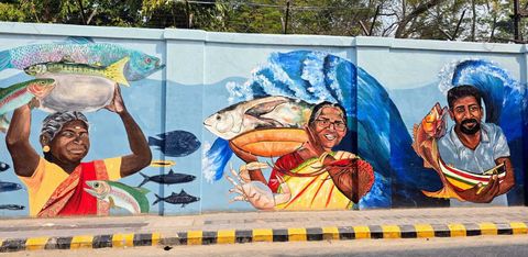 A street wall mural showing fisherwomen and a fisherman carrying large fish, with a blue ocean wave painted behind them, part of the Guardians of Fort Kochi mural.