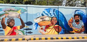 A street wall mural showing fisherwomen and a fisherman carrying large fish, with a blue ocean wave painted behind them, part of the Guardians of Fort Kochi mural.