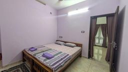 Bedroom with lilac walls, a wooden double bed with striped purple and white bedding and folded purple towels, a ceiling light strip and an open door to a room beyond.