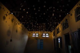 Hundreds of small glass baubles hanging in rows from the dark timber roof of a tall room, catching the light above pale walls and arched windows.