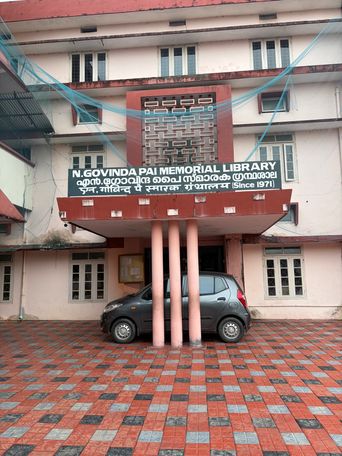 A multi-story building with a paved courtyard in front, where a dark car is parked beneath a covered entrance supported by thick columns. A large sign is mounted above the entrance, and blue netting is strung across the upper facade. The building has multiple windows and a decorative lattice panel above the sign.