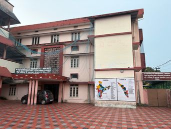 A multi-story building with a paved courtyard in front, where a dark car is parked under a covered entrance supported by pillars. On the side of the building, two large maps are mounted side by side, one showing India and the other showing Kerala. A signboard is visible above the entrance, and another signboard is mounted on a wall to the right. The building has multiple windows and a flat roofline with red trim.