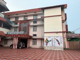 A multi-story building with a paved courtyard in front, where a dark car is parked under a covered entrance supported by pillars. On the side of the building, two large maps are mounted side by side, one showing India and the other showing Kerala. A signboard is visible above the entrance, and another signboard is mounted on a wall to the right. The building has multiple windows and a flat roofline with red trim.