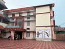 A multi-story building with a paved courtyard in front, where a dark car is parked under a covered entrance supported by pillars. On the side of the building, two large maps are mounted side by side, one showing India and the other showing Kerala. A signboard is visible above the entrance, and another signboard is mounted on a wall to the right. The building has multiple windows and a flat roofline with red trim.