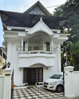 White house with a pointed gable, a railed balcony above an arched porch, and a white car parked on a checkered tile drive.