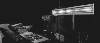 A dark room with a DJ deck and mixer on a table, a speaker stack behind, and a long strip light hanging overhead.