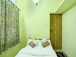 Small pale green bedroom with a white double bed, two patterned cushions, a leaf-print curtain on the left and a wooden door on the right.