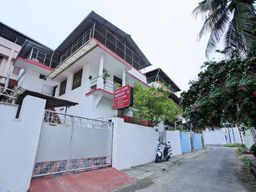 Two-storey white house behind a white gate, with a red sign board on its front wall, a balcony with a railing and a parked scooter in the lane beside it.