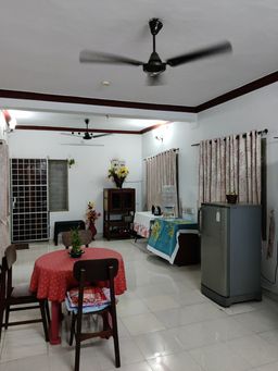 Open room with a round dining table under a red cloth, a grey fridge, a water dispenser on a side table, and curtained windows.