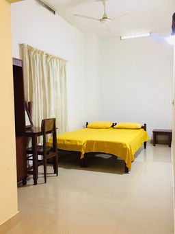 Bedroom with two beds pushed together under a yellow bedspread, a wooden dressing table with a mirror and chair at the side, a cream curtain and a ceiling fan.