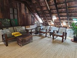 A loft-style room under an exposed timber-and-tile roof, with a cane-backed wooden sofa and armchairs on a woven mat, a wooden chest, a standing fan, and a potted plant at the right.