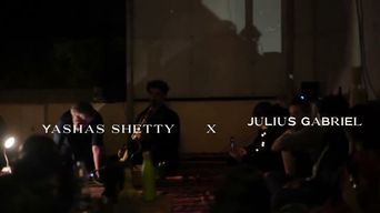 A dim video still from a night performance, an audience seated on mats around two musicians, with the words YASHAS SHETTY X JULIUS GABRIEL across the frame.