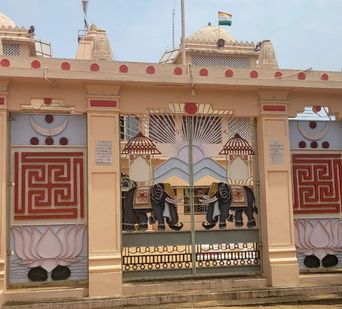 A decorative gate with two painted elephants facing each other beneath a sunburst design, flanked by pillars with geometric and lotus motifs, topped by a building with domes and a flag flying above.