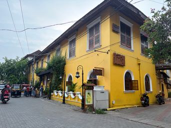 A bright yellow two-story building stands at a street corner, featuring arched windows on the ground floor and rectangular windows above, with several wooden signs mounted on its facade. A low white wall with decorative elements runs along the front, beside a paved sidewalk where a few people are gathered near an auto-rickshaw. A motorcyclist rides past on the road, while parked motorcycles are visible along the building’s side. Overhead wires cross the sky, and green trees frame the scene on both sides.
