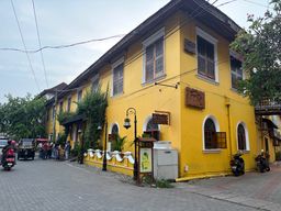 A bright yellow two-story building stands at a street corner, featuring arched windows on the ground floor and rectangular windows above, with several wooden signs mounted on its facade. A low white wall with decorative elements runs along the front, beside a paved sidewalk where a few people are gathered near an auto-rickshaw. A motorcyclist rides past on the road, while parked motorcycles are visible along the building’s side. Overhead wires cross the sky, and green trees frame the scene on both sides.