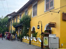 A bright yellow building with white-trimmed windows and arched doorways lines a paved street, with climbing greenery covering part of its facade. A wooden sign hangs from the wall above a small wooden stand holding a poster, next to a white utility box. Black lampposts with glass lanterns stand along a low white wall with scalloped edges. Several people are walking on the street and gathered near the entrance of the building, which has a covered walkway supported by dark pillars. Overhead wires run across the sky above the building.