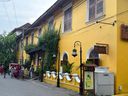 A bright yellow building with white-trimmed windows and arched doorways lines a paved street, with climbing greenery covering part of its facade. A wooden sign hangs from the wall above a small wooden stand holding a poster, next to a white utility box. Black lampposts with glass lanterns stand along a low white wall with scalloped edges. Several people are walking on the street and gathered near the entrance of the building, which has a covered walkway supported by dark pillars. Overhead wires run across the sky above the building.