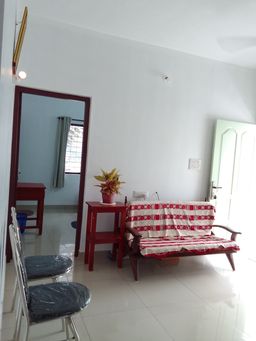 Living room with white walls and a glossy tiled floor, a wooden sofa with a red-and-white cover, a red side table with a potted plant, and chairs in clear plastic covers.