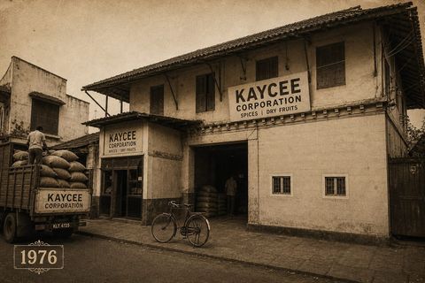 The Kaycee Corporation warehouse in Mattancherry in 1976. A painted board above the open loading bay reads KAYCEE CORPORATION, SPICES | DRY FRUITS. A man stacks sacks onto a truck at the kerb and a bicycle leans by the door.