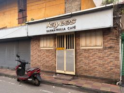 A red scooter is parked on the street in front of a brick building with a white sign above the entrance. The entrance has a metal gate with a yellow notice attached, flanked by two closed wooden shutters. To the left of the entrance is a closed metal roller shutter. The building has a yellow upper wall with a window and visible overhead wires.