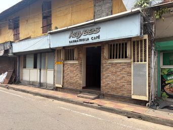 A street-level view of a single-story building with a brick facade and a light blue sign above the entrance. The entrance is an open doorway leading into a dark interior, flanked by windows with wooden bars. To the left of the entrance is a closed storefront with glass panels and a metal shutter. To the right, a narrow vertical panel with a mesh screen stands beside the doorway. The building sits on a paved sidewalk next to an asphalt road. Overhead electrical wires run across the front of the building, and a green storefront with a poster is visible on the far right edge.