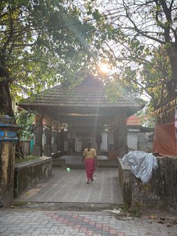 A person wearing a yellow top and a red lower garment walks away from the camera along a paved path toward a traditional building with a tiled roof and open front. The sun shines through tree branches above, casting light across the scene. Stone walls line the path on both sides, with a covered object resting on the right wall and a small green object on the ground nearby. Trees frame the structure, and a string of triangular flags hangs across the front of the building.