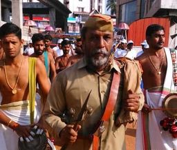 A man in a khaki uniform and cap holding a ceremonial item, walking in a procession among men in traditional white and gold dress.