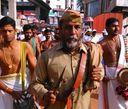A man in a khaki uniform and cap holding a ceremonial item, walking in a procession among men in traditional white and gold dress.