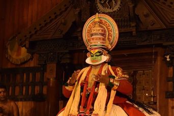 A Kathakali performer in a tall crown, with the painted green face and white frame of the pacha make-up, seated on a carved wooden stage.