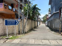 A paved street stretches between a textured wall on the left and a building wall on the right, with a blue directional sign mounted on two posts beside the left wall. Above the wall, laundry hangs from a balcony of an orange building, and banana trees grow behind utility poles and wires. A scooter is parked along the right side of the road, which recedes into the distance under a clear sky.