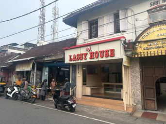 A street-level view of a row of shops under an overcast sky, with overhead power lines and communication towers visible in the background. The central shop has a red sign above its entrance and a glass-fronted interior with shelves and a counter. Several people stand near parked scooters on the road in front of the shops, while another person stands inside the central shop’s doorway. To the right, an adjacent building features an arched entrance with a wooden door and a decorative sign above it.