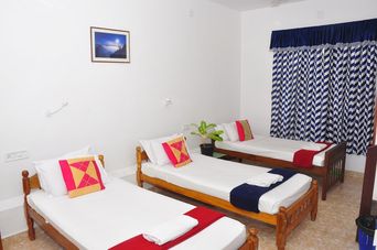 Room with three single wooden beds with white bedding and red-and-gold cushions, blue-and-white striped curtains, and a framed picture on the wall.