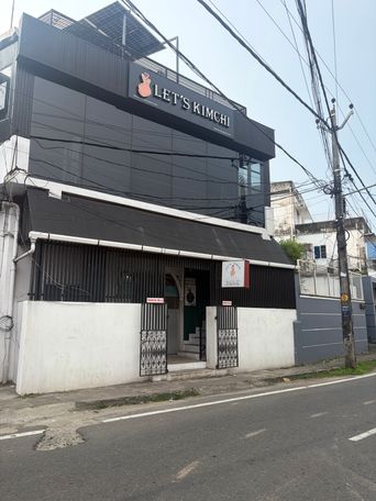 A two-story building with a black facade stands on a street corner, featuring a sign with a logo above the entrance. The ground floor has a covered entryway with black metal gates on either side, leading to a doorway and a short flight of stairs. A white wall runs along the front, and a smaller sign is mounted near the entrance. Overhead, numerous electrical wires crisscross the sky above a utility pole on the right. The road in the foreground has white painted lines.