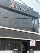 A building with a dark facade and large glass windows on the upper level, featuring a sign with a hand gesture icon and text above the entrance. Below, a covered walkway with vertical slats leads to a doorway, with a smaller sign mounted on the wall to the right. Several small signs are visible near the ground level, and overhead wires cross in front of the building.