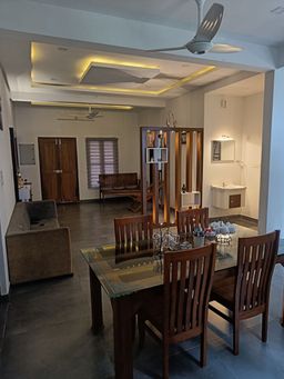 Open-plan living and dining room with a glass-topped table and four wooden chairs, a grey sofa at the side, dark wooden doors and a ceiling fan under lit recessed panels.
