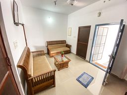 Living room with two wooden sofas with tan cushions around a glass-top coffee table, a blue rug on a tiled floor, and an open door to outside.