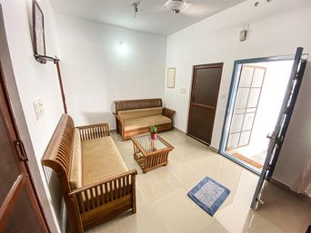 Living room with two wooden sofas with tan cushions around a glass-top coffee table, a blue rug on a tiled floor, and an open door to outside.