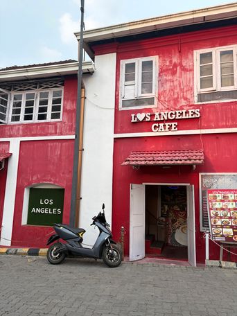 A red building with white trim houses a cafe, with its name displayed above the entrance and on a green panel on the side wall. A grey scooter is parked on the paved street in front of the open doorway, where a staircase is visible inside. A menu board stands on the right side of the entrance, and a tall pole rises beside the building.