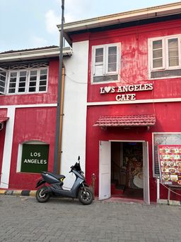 A red building with white trim houses a cafe, with its name displayed above the entrance and on a green panel on the side wall. A grey scooter is parked on the paved street in front of the open doorway, where a staircase is visible inside. A menu board stands on the right side of the entrance, and a tall pole rises beside the building.