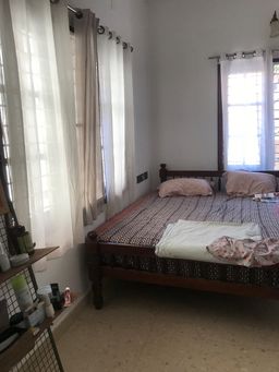 Bedroom with a wooden bed under a patterned bedspread, sheer white curtains at two windows, and a shelf of toiletries and bottles at the left.