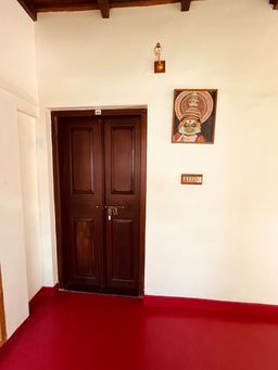 A corridor with a pair of dark wooden double doors set in a cream wall, a framed Kathakali mask painting beside them, and a red-painted floor.