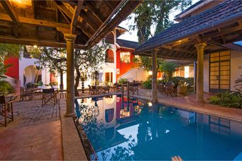 Courtyard pool at dusk ringed by tiled-roof verandas on stone pillars, with tables and chairs and red and white walls behind.