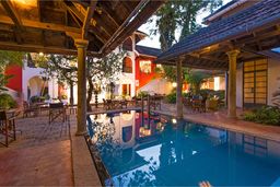 Courtyard pool at dusk ringed by tiled-roof verandas on stone pillars, with tables and chairs and red and white walls behind.