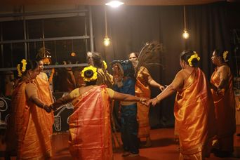 Women in orange and gold saris with yellow flowers in their hair hold hands in a circle around a costumed figure carrying peacock feathers, under hanging filament bulbs.