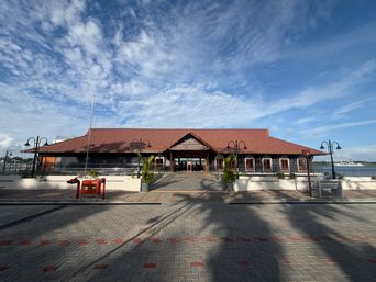 A long building with a red tiled roof stands under a blue sky filled with scattered white clouds. The structure has a central entrance flanked by windows and is approached by a paved walkway. Several tall lampposts line the front, and potted plants sit near the entrance. In the foreground, a paved area with red markings stretches toward the building, and a red object is visible on the left side. A body of water can be seen in the distance behind the building.